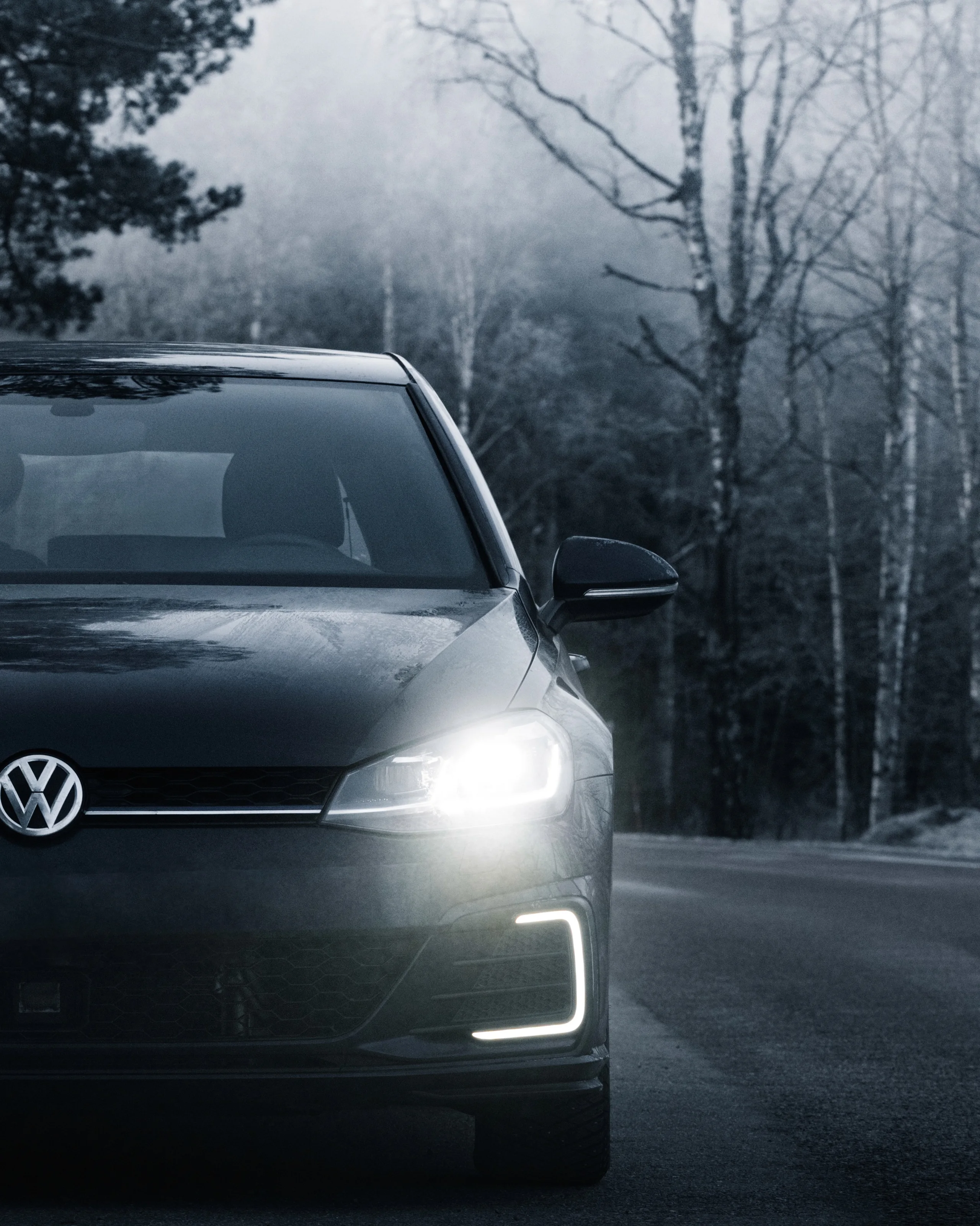 Dark Volkswagen hatchback with headlights on, on a misty forest road