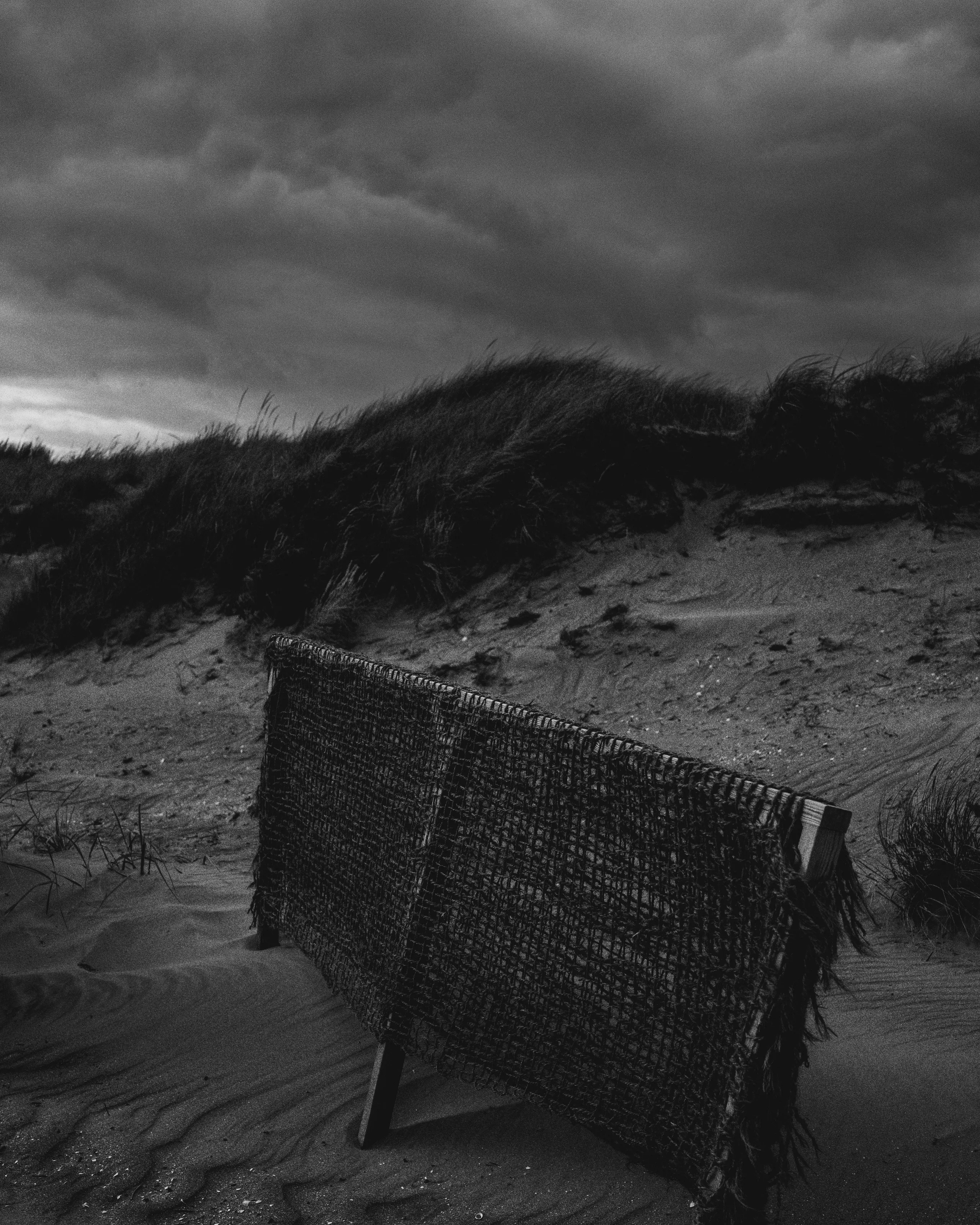 Black and white image of a weathered fence half buried in sand dunes